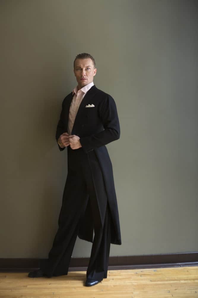 A man wearing frock coat stands against the wall.