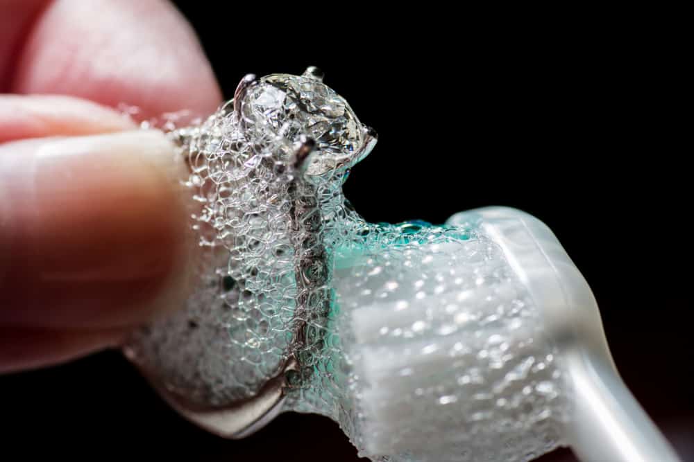 Woman's hands cleaning a diamond ring with toothbrush and soap.