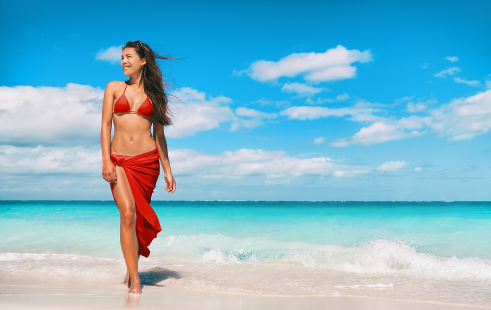 Model in red swimsuit and sarong poses by the beach.