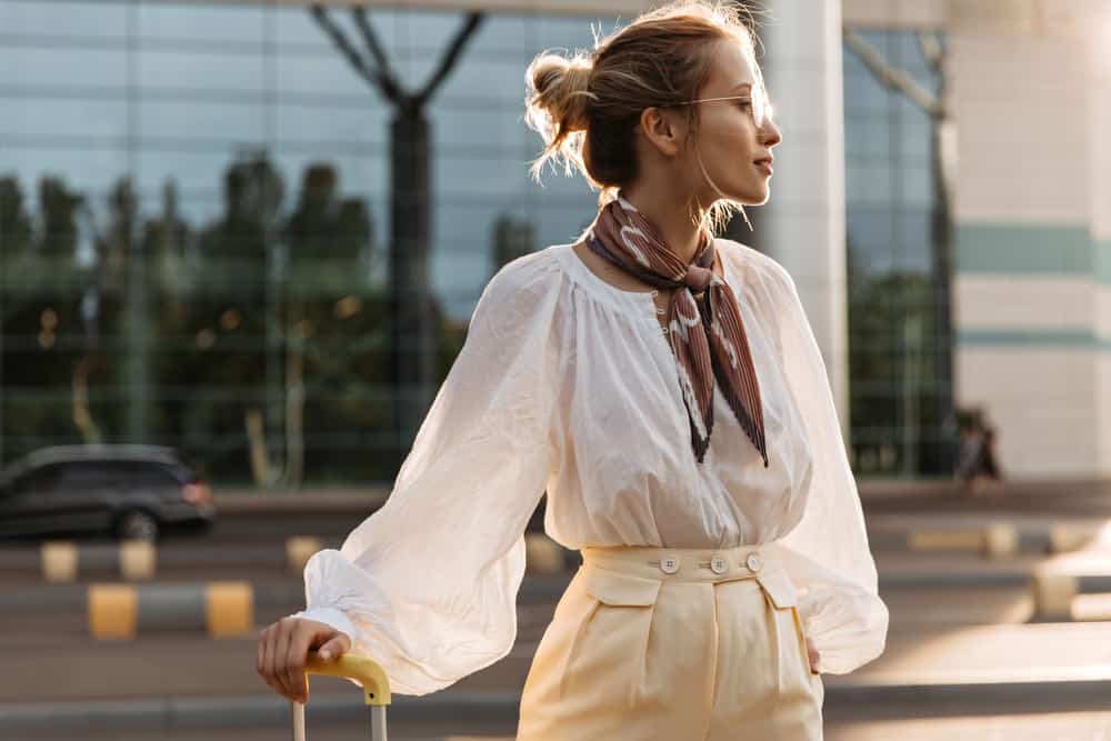 Portrait of blonde girl in white blouse, beige pants, brown silk scarf and eyeglasses holding luggage.