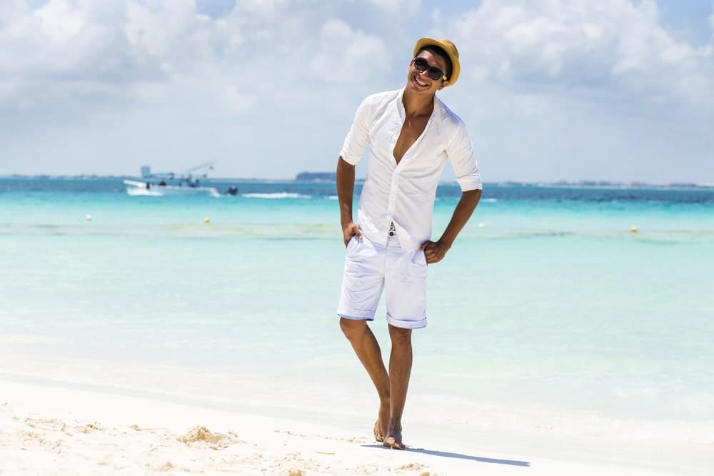 A man wearing neutral tones with his hat and sunglasses at the beach.