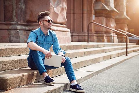 Man in denim polo and pants sitting on stairs outside the building.