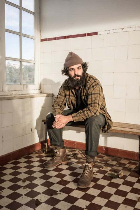 Bearded man in rugged polo and pants sitting on a bench.