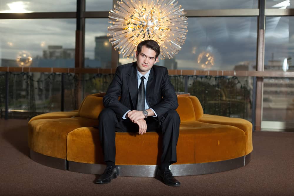 Man in black suit and tie sitting on a luxury chair.