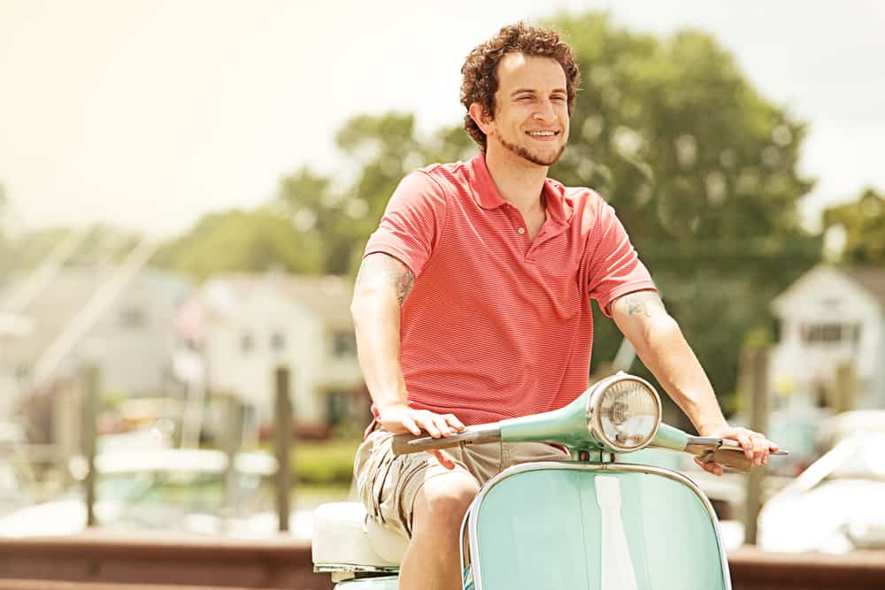 This is a man wearing a khaki cargo shorts with a salmon polo shirt while riding a scooter.