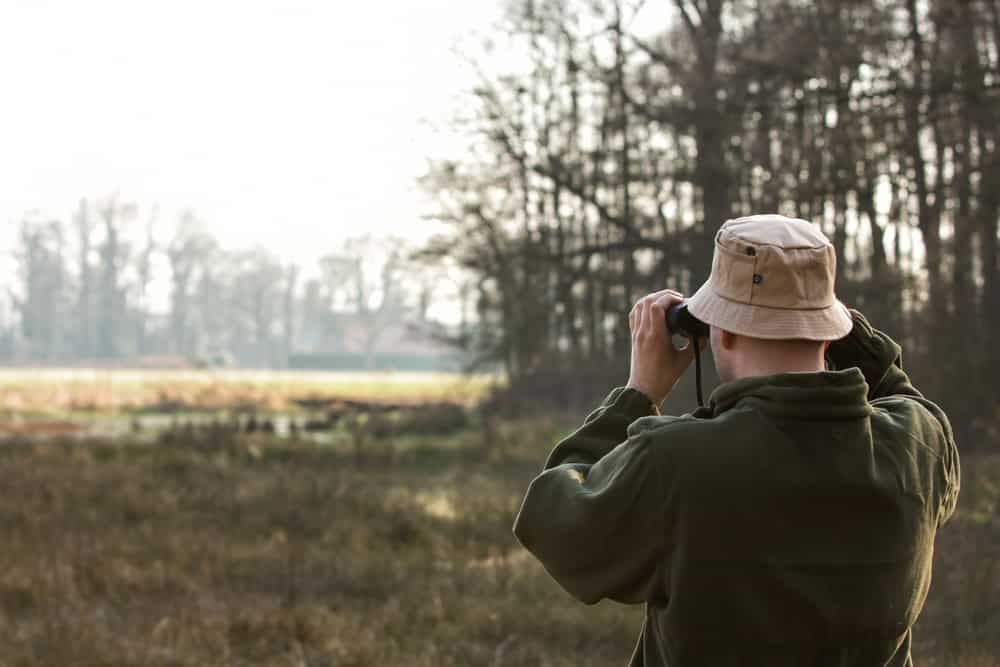 A bird watcher wearing a hat and a coat using his binoculars.