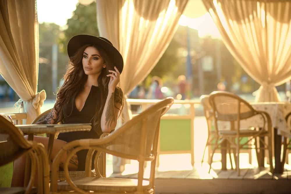 This is a woman staying at the al fresco section of the restaurant wearing a fedora.