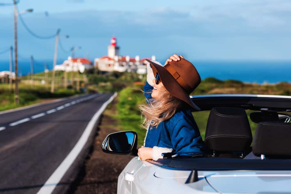 A woman riding in a convertible car holding onto her hat.