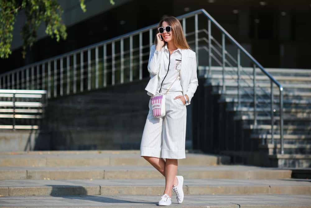 Woman wearing a matching jacket and bermuda shorts walks through the city while engaged in a call.