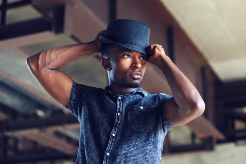 A man wearing a dark denim shirt with his dark short-brimmed fedora.