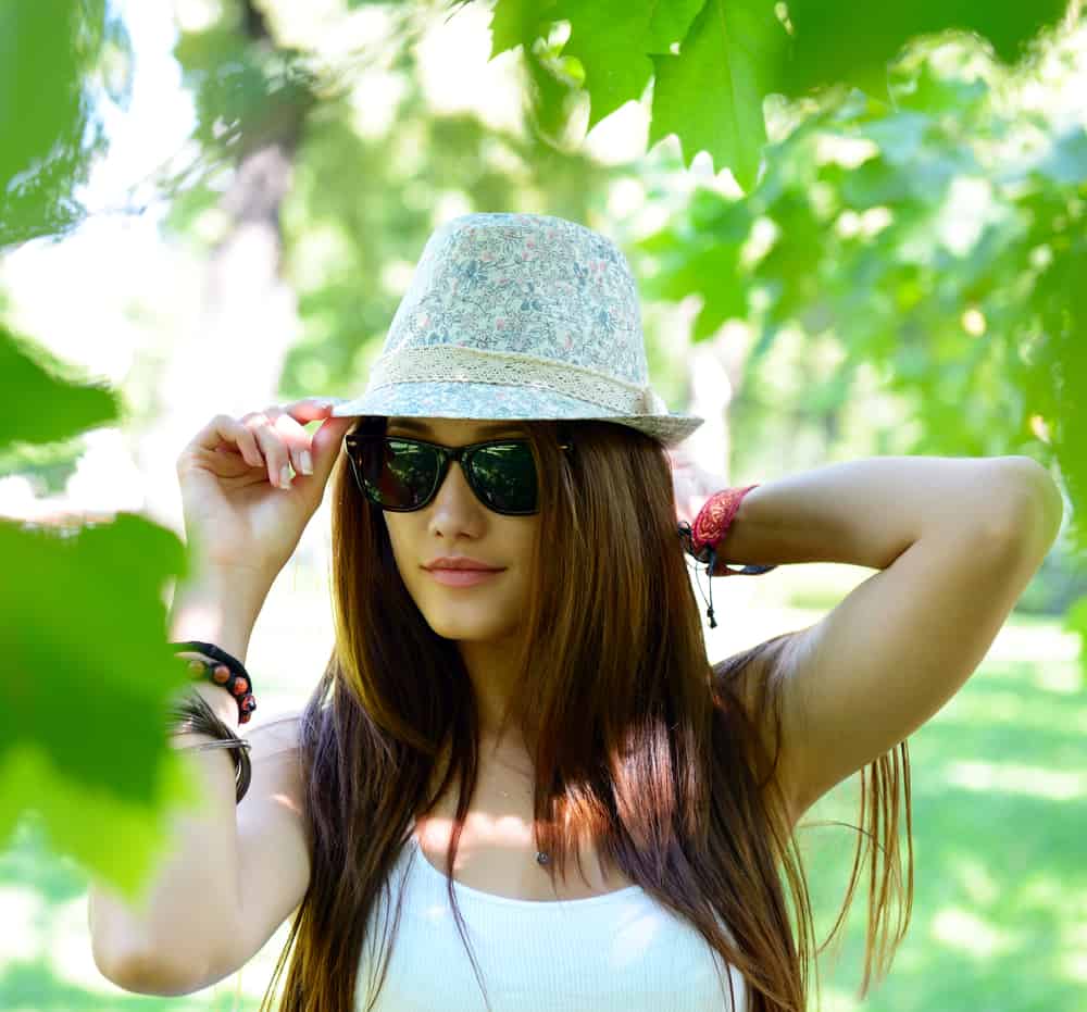 A woman wearing a white tank top, sunglasses and a pastel-toned patterned fedora.